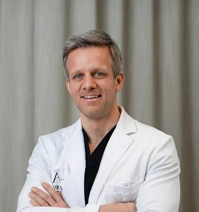 Dr. Jes Rødgaard fra Asteta Clinic, iført hvit labfrakk og smilende med armene i kors foran et nøytralt bakteppe.