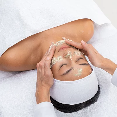 Kvinne får ansiktsbehandling, liggende på hvitt håndkle, med green peel ansiktsmaske påført av behandlerens hender.