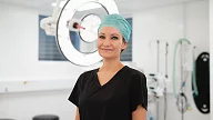 Person smiler, står i operasjonsrom foran medisinsk utstyr, iført kirurgisk antrekk.