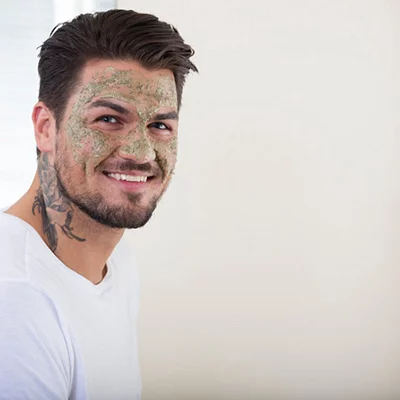 Manne med ansiktsmaske og tatovering, smilende mens han ser til siden. Han har på seg en hvit t-skjorte.