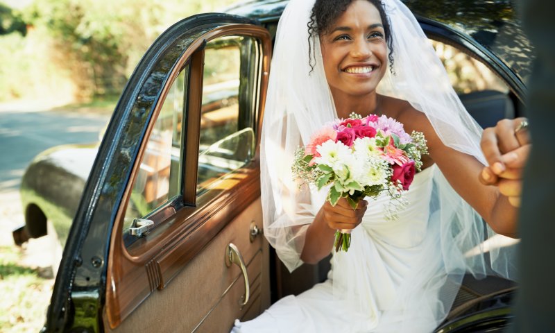 Smilende brud med blomsterbukett hjelpes ut av bil